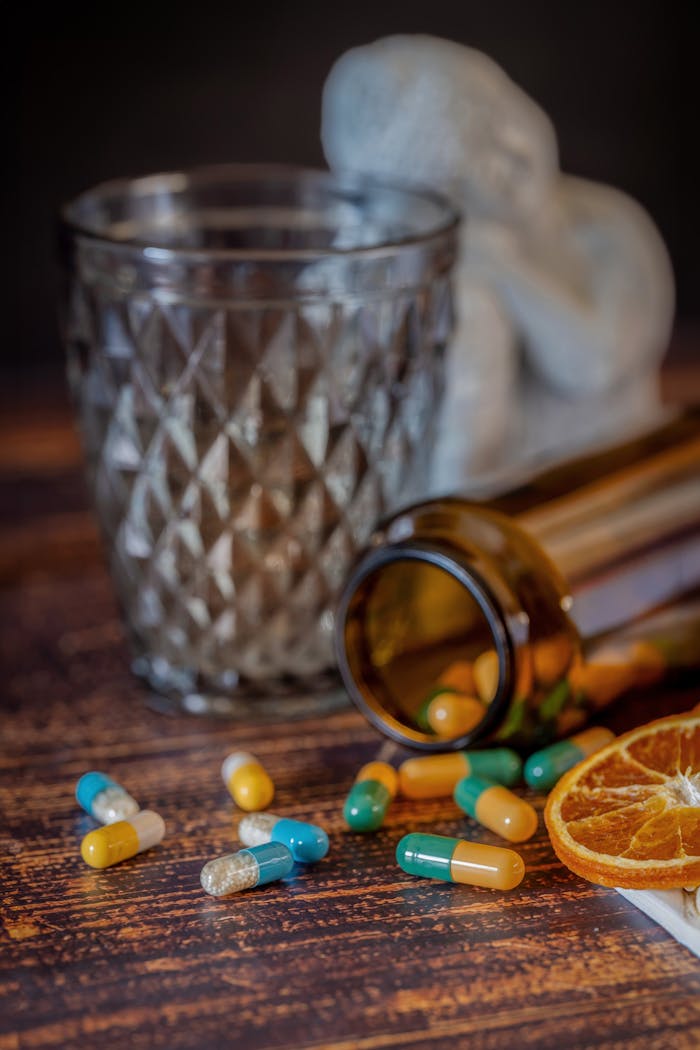 Colorful pills and orange slices on a wooden table with a glass and bottle in the background.