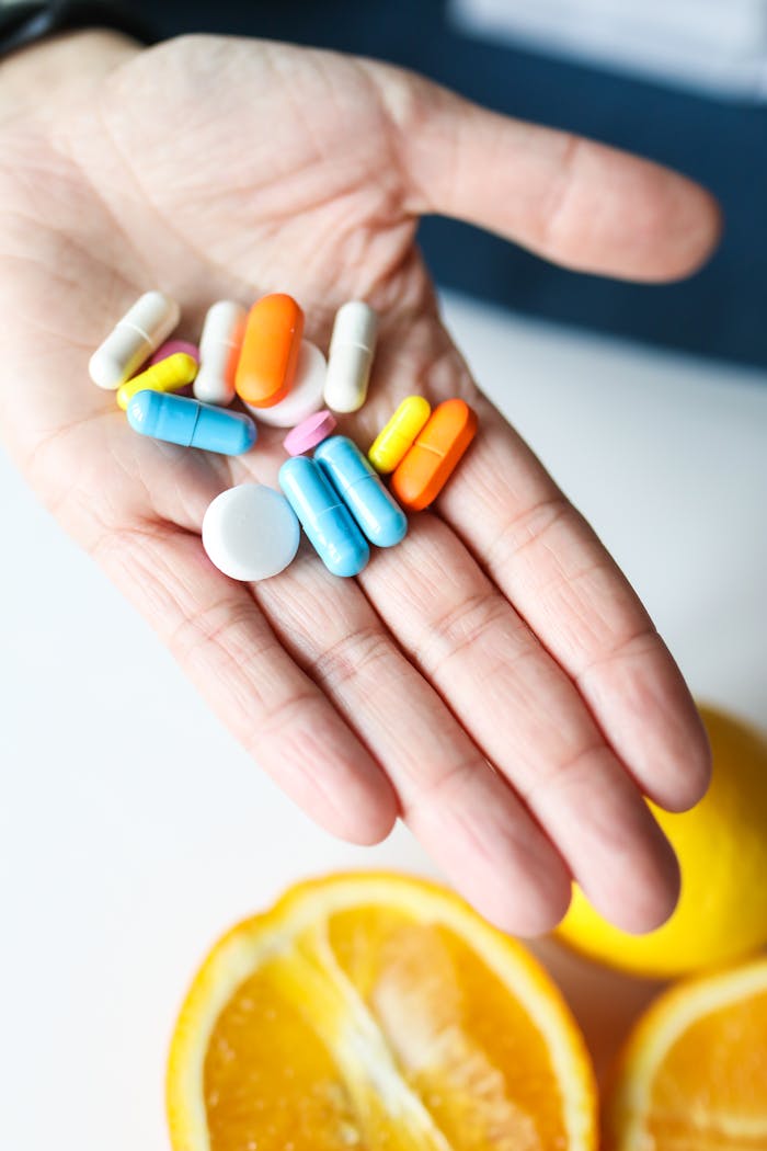 Hand holding colorful pills next to fresh orange slices, promoting health and vitamin intake.